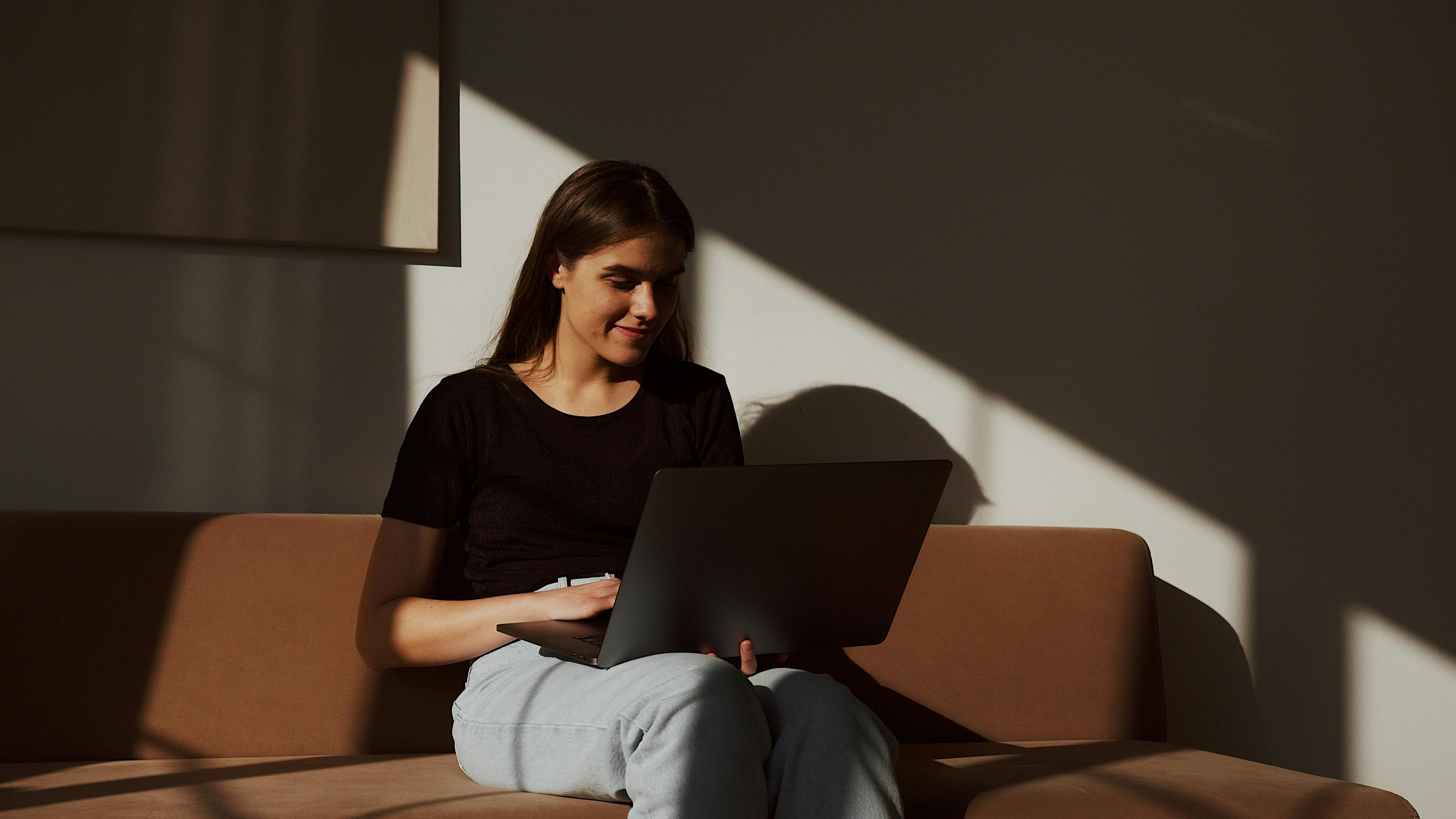 cheerful woman using laptop on sofa in shadows