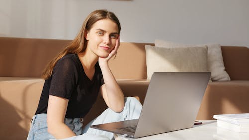 Positive glad young lady in casual clothes sitting on floor and browsing modern netbook while leaning head on hand and looking at camera happily