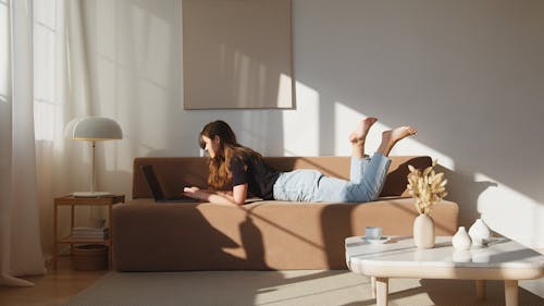 Free Side view full body young lady in casual outfit lying on comfortable couch and browsing netbook in cozy modern living room Stock Photo