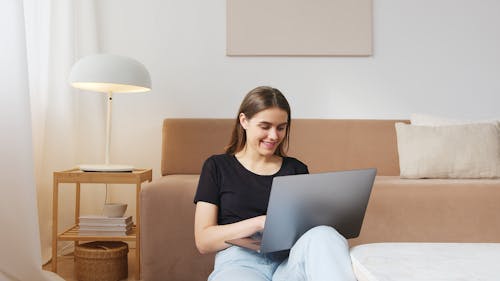 Smiling young lady in casual wear browsing modern laptop and sitting on floor near comfy sofa in light living room