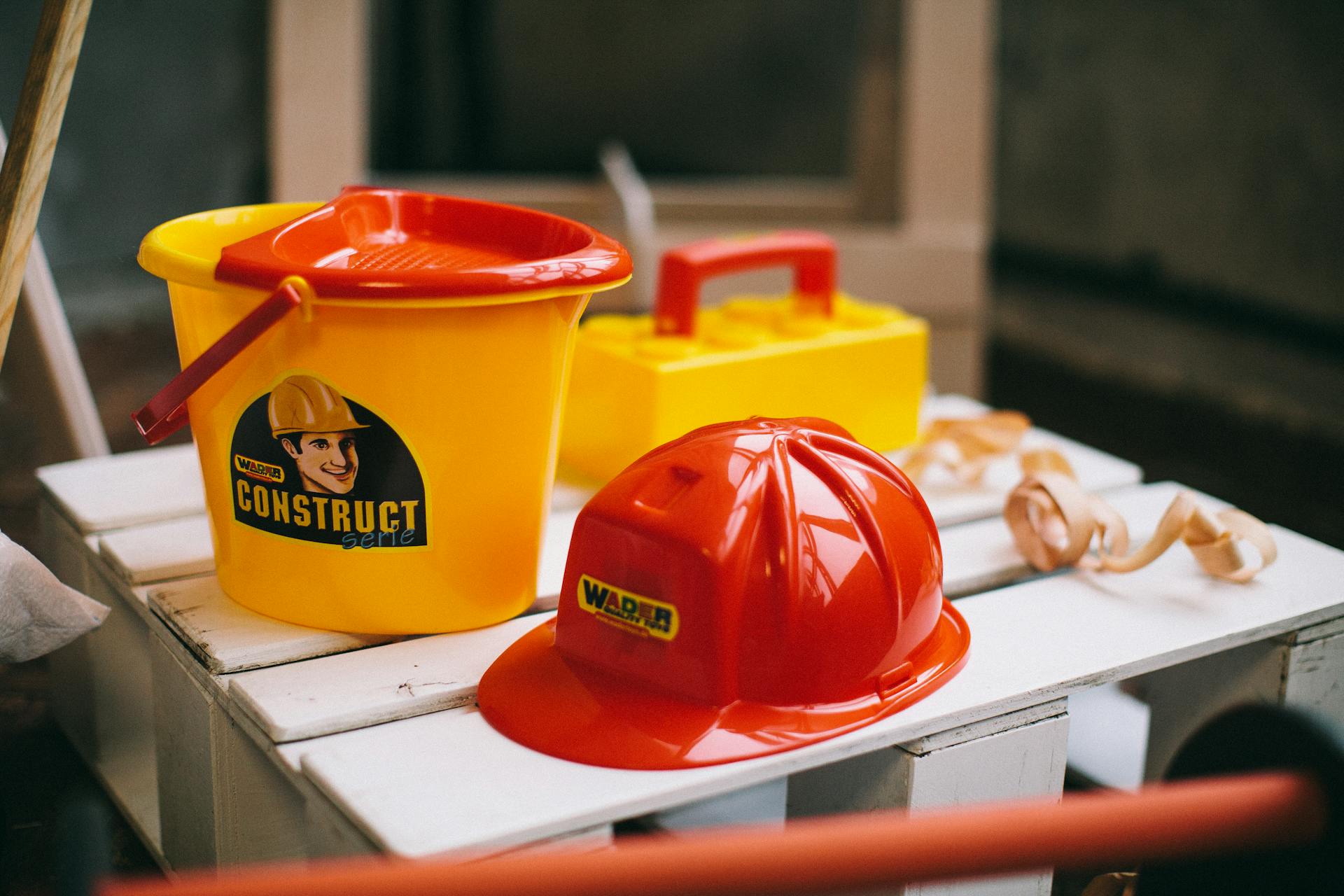 A Red Plastic Hard Hat Toy and a Yellow Bucket on a Wooden Crate