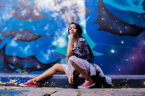 Free Young ethnic female with dark hair in casual clothes sitting on curb against wall with blue graffiti Stock Photo