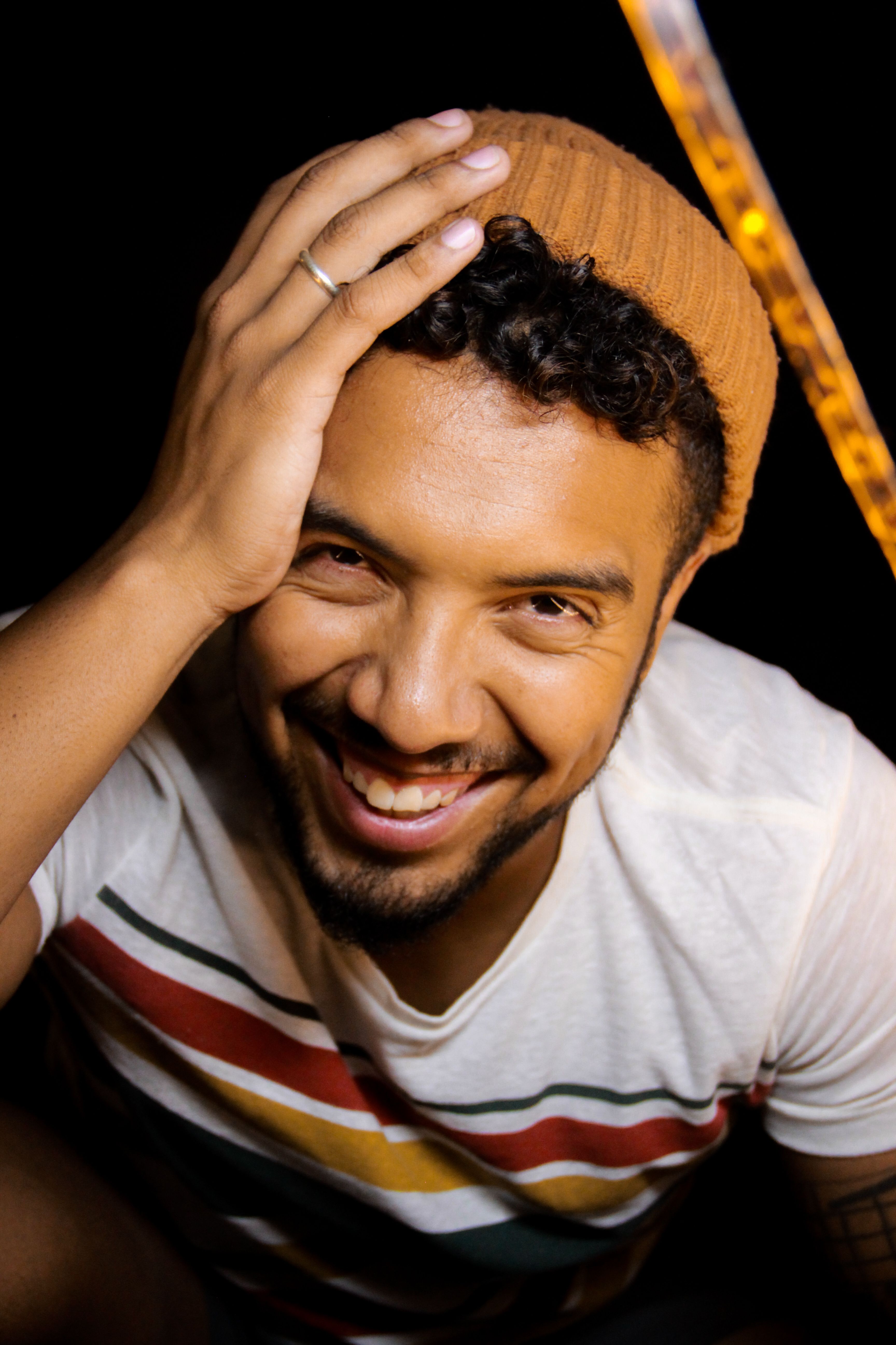 Happy young ethnic male with curly hair and beard in casual clothing and warm beige hat touching head and looking at camera with bright smile