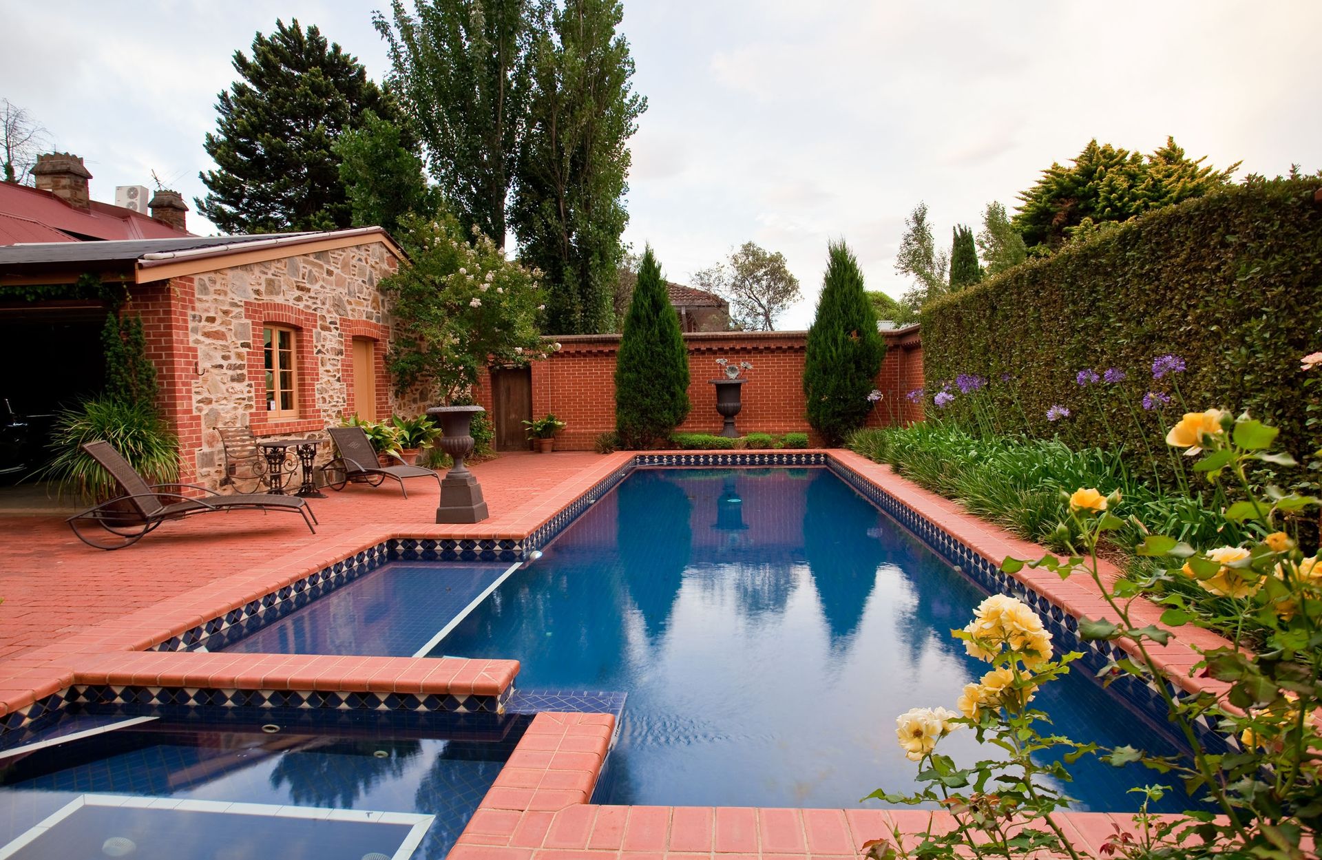 Big blue swimming pool in yard of small brick private house decorated with tall green hedgerows and blooming flowers