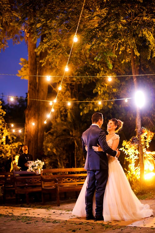 Free Full body of young married couple in elegant wedding dress looking at each other while dancing in garden next to wooden benches under glowing garlands Stock Photo