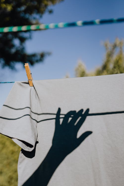 Free Clean tee shirt with shadow of hand of faceless woman drying on sun on backyard during cloudless sunny day Stock Photo