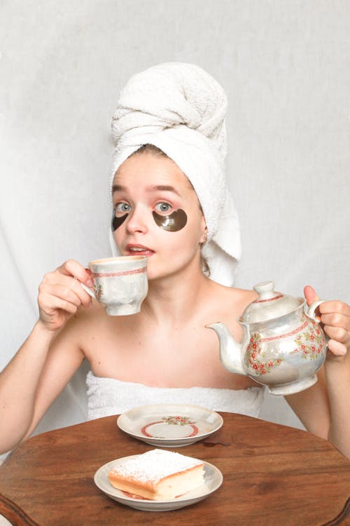 Surprised young female in towel on chest and head and patches on face looking at camera while holding teapot and drinking beverage at small round wooden table in bright room