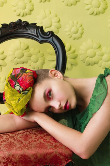 Serious stylish female in green dress and headscarf with hair tied up looking at camera while lying with head on soft seat of chair in bright room