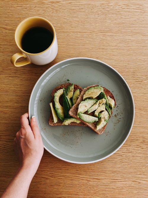 Free Overhead view of unrecognizable person holding big round plate with sandwich with avocado near cup of tea on light brown wooden table in bright room Stock Photo