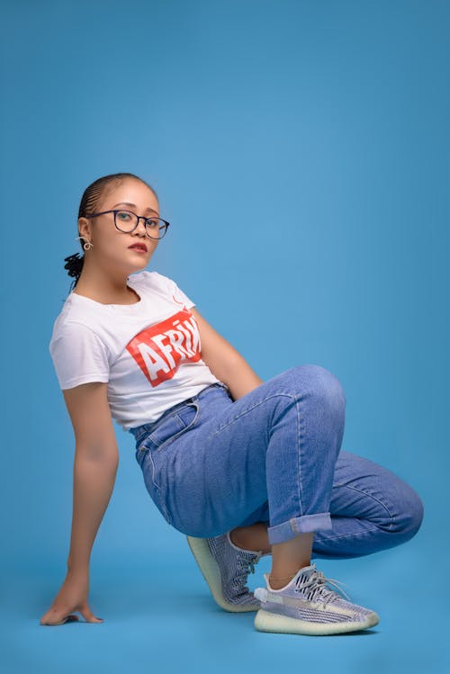 Full body of young ethnic female with cornrows in casual clothing and eyeglasses looking at camera while squatting in bright studio on blue background