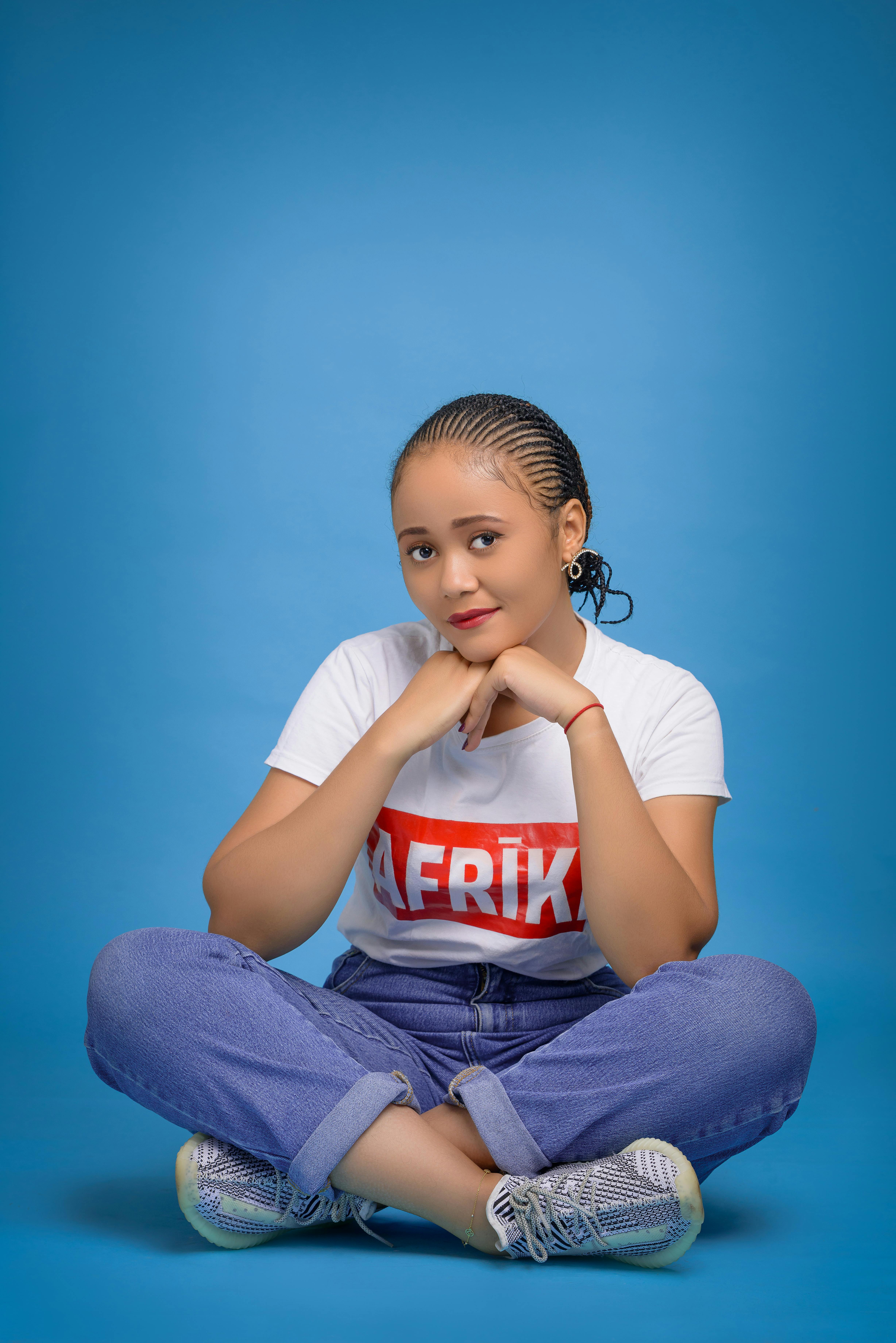 Full body of young ethnic female in shirt and jeans looking at camera while chilling on floor with legs crossed and hands under chin on blue background