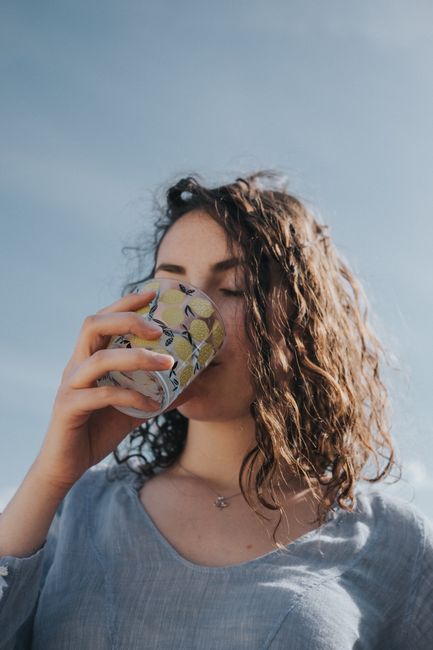 A young woman with wavy hair enjoys a refreshing drink from a colorful glass under a clear blue sky.