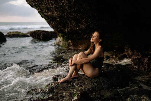 Side view of young female in swimsuit relaxing and dreaming while sitting and hugging knees on rocky shore near sea