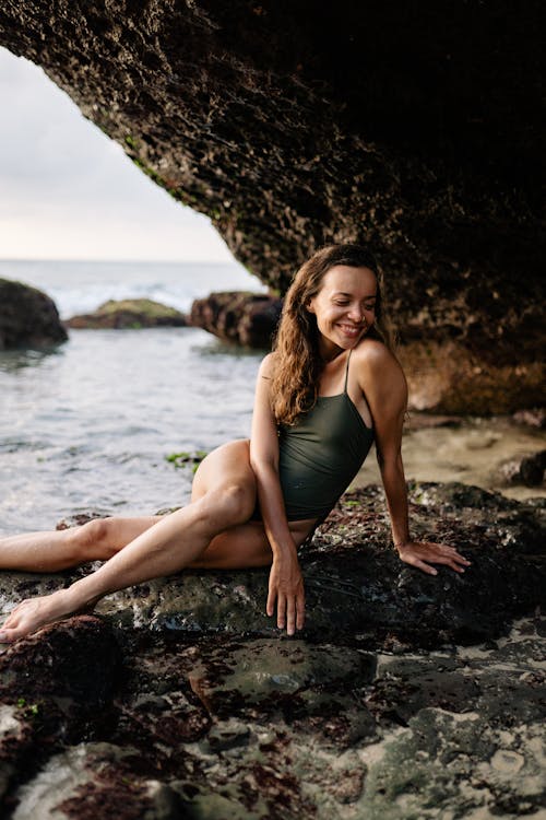 Happy young woman in swimsuit smiling and enjoying rest while sitting on stone and leaning on hand against sea on daytime
