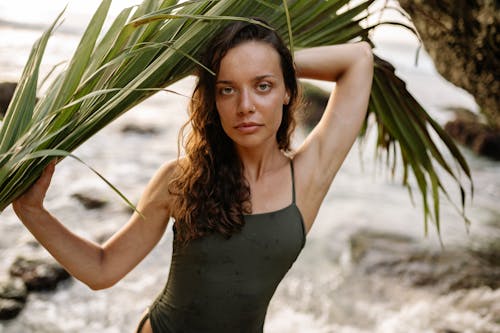 Young good looking female model in swimming suit with green palm leaf in hands looking at camera while relaxing on sunny beach