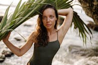 年轻好看的女模特穿着泳衣，手里拿着绿色的棕榈叶，在阳光明媚的海滩上放松时看着相机