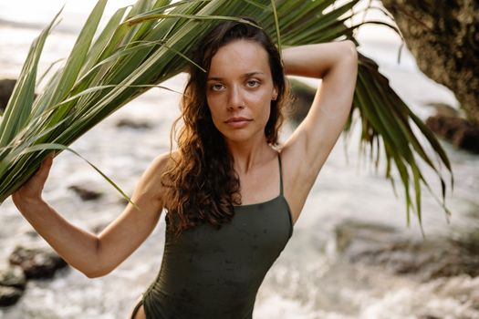 一位年轻英俊的女模特穿着泳衣，手持绿色棕榈叶，在阳光明媚的海滩上放松身心，注视着镜头