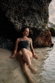 Full body emotionless female in swimwear relaxing alone on wet sand in sea water while leaning on rocky cliff in sunny evening