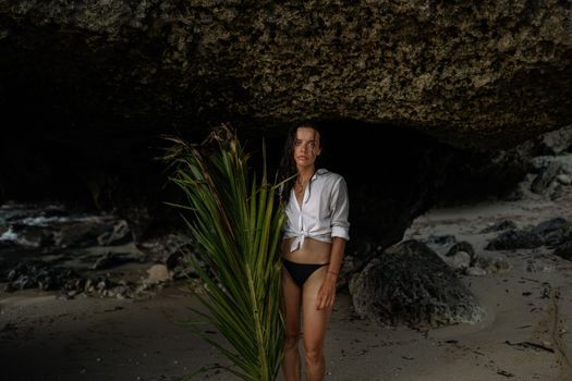一名妇女站在热带海滩的岩石掩体下，四周棕榈叶环绕，环境宁静