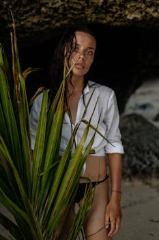 一位冷酷无情的年轻女旅行者，穿着比基尼、外系衬衫，手持棕榈枝站在山洞中