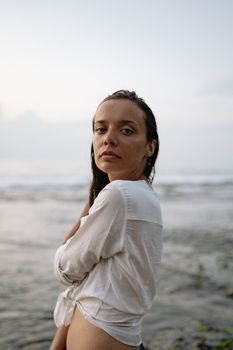 Side view of calm female in white tied shirt standing on coast against sea water under overcast sky