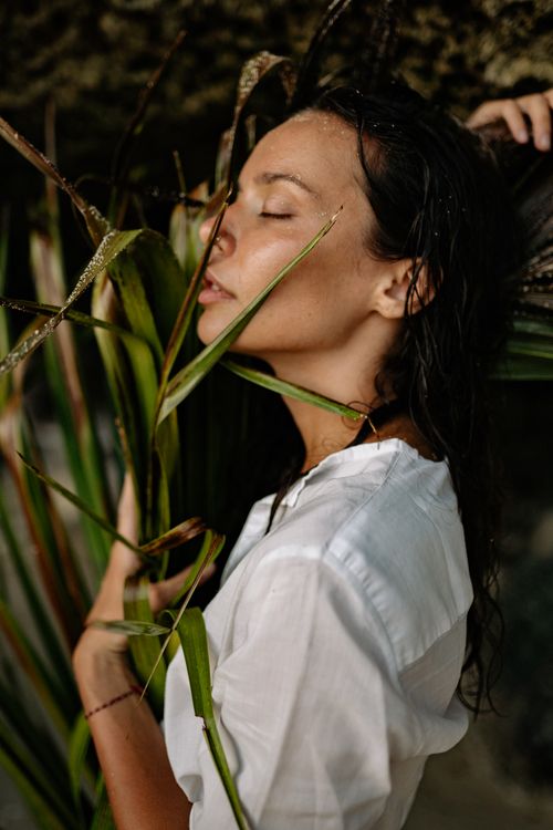 Free Side view of young female with closed eyes in white clothes cuddling branch of palm to chest while enjoying nature on beach Stock Photo