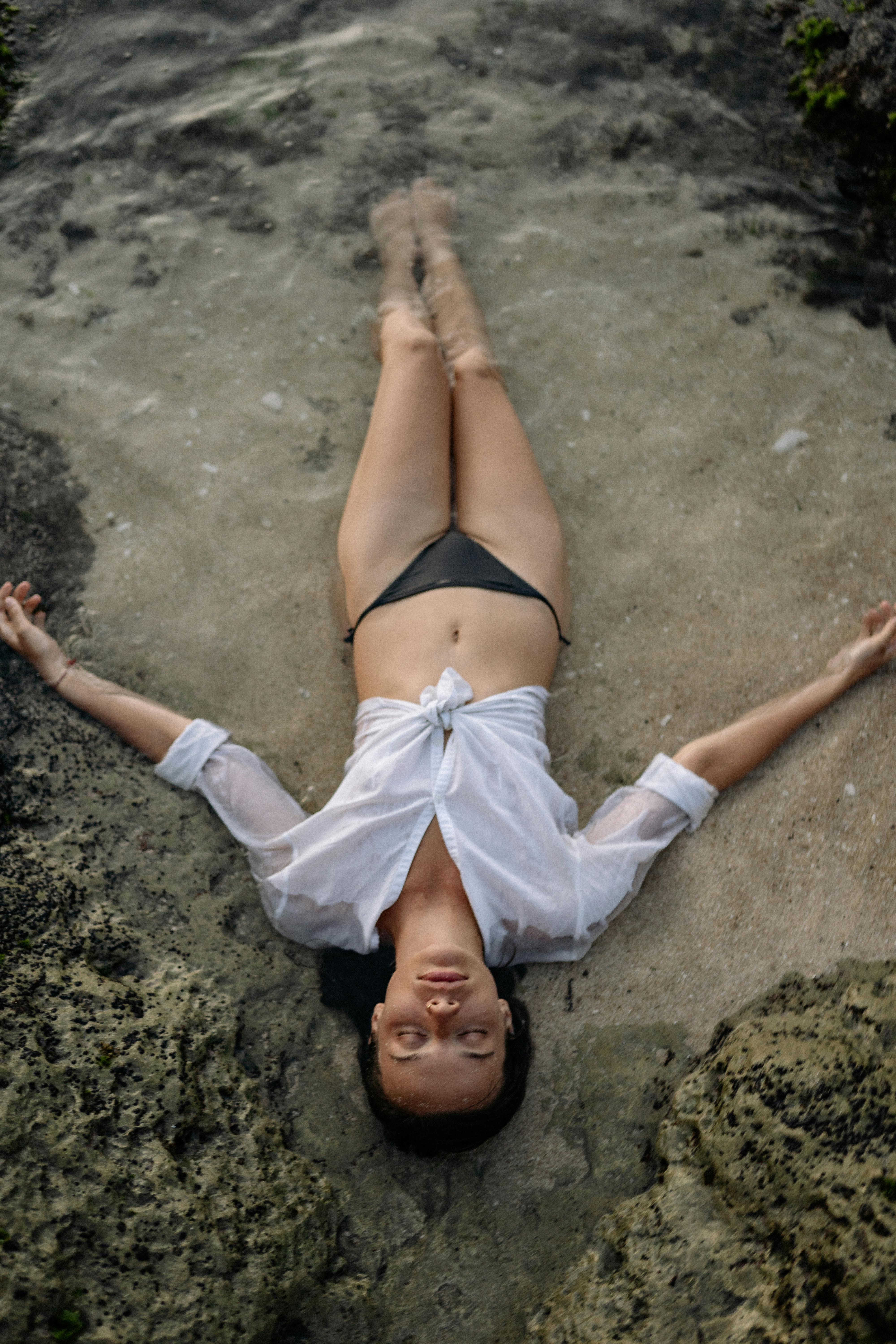 From above of full length slim female with closed eyes in white shirt and black bikini lying on sandy shore in water while enjoying time