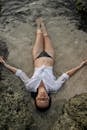 From above of full length slim female with closed eyes in white shirt and black bikini lying on sandy shore in water while enjoying time