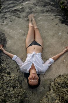 从上方看去，一位身材修长、身着白衬衫和黑色比基尼的女性正闭着眼睛躺在水边的沙滩上享受时光