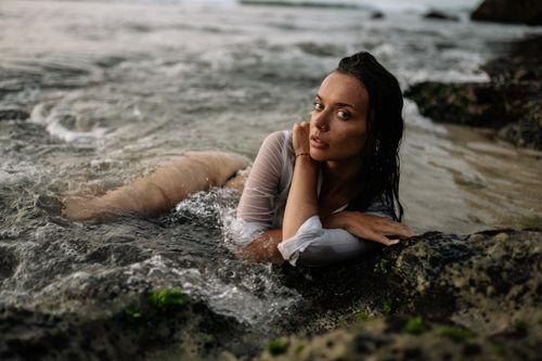 Free Full body of slim young female in white shirt and black bikini lying with crossed arms on sandy coast while sea water waving and covering body of woman Stock Photo