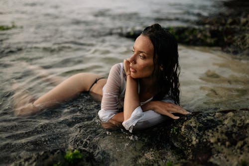 Full body slim female in white shirt and black bikini enjoying vacation while lying alone in sea water on coast