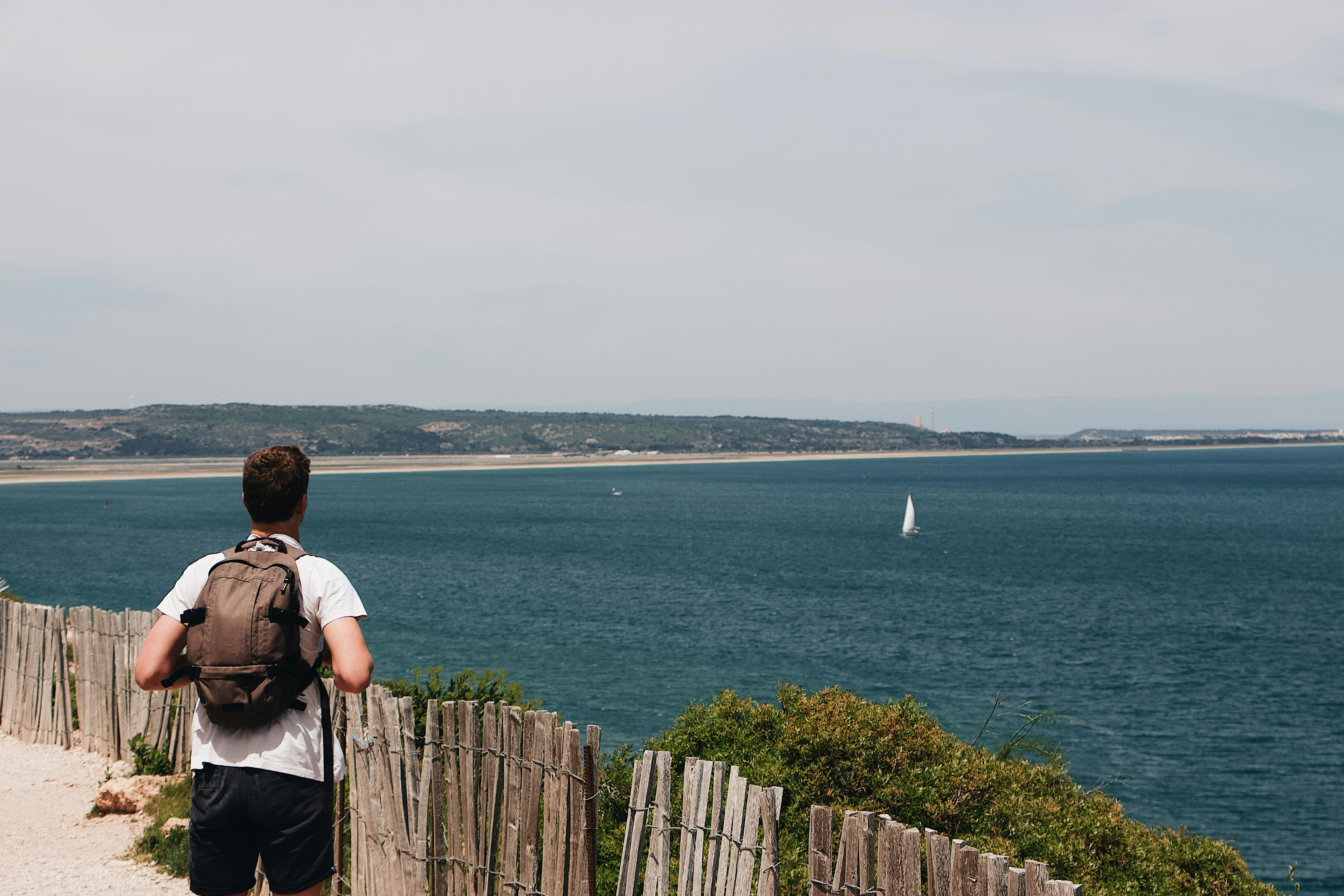 A man with a backpack stands by a coastal fence in Leucate, France, overlooking the serene sea.