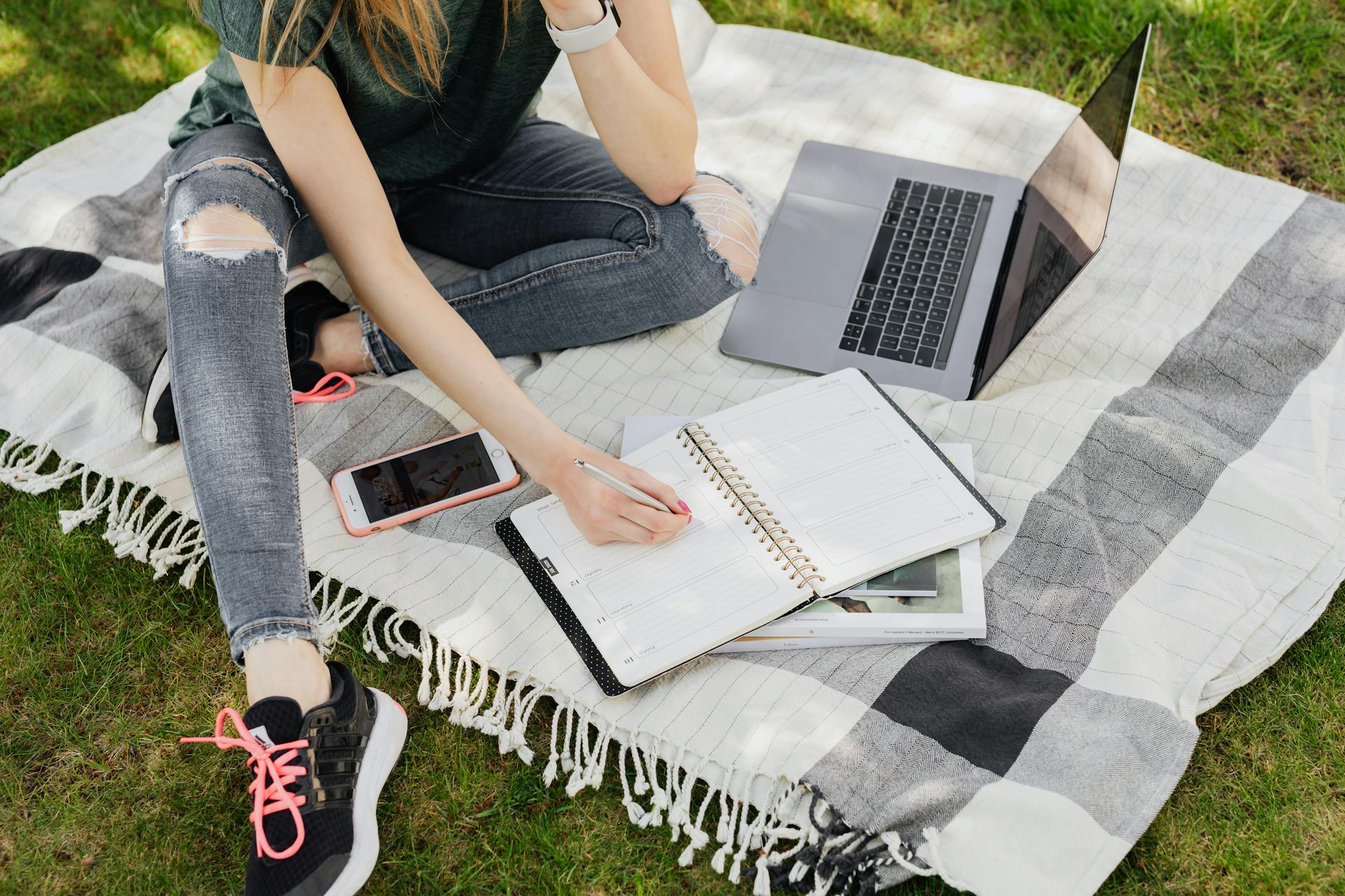 From above crop anonymous female student in casual outfit writing in planner while sitting on warm blanket with smartphone and laptop on green park lawn