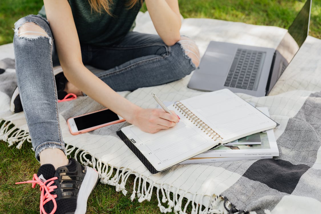 Free Crop faceless female student in casual clothes taking notes in planner while sitting on blanket with smartphone and laptop on green park grass on sunny day Stock Photo