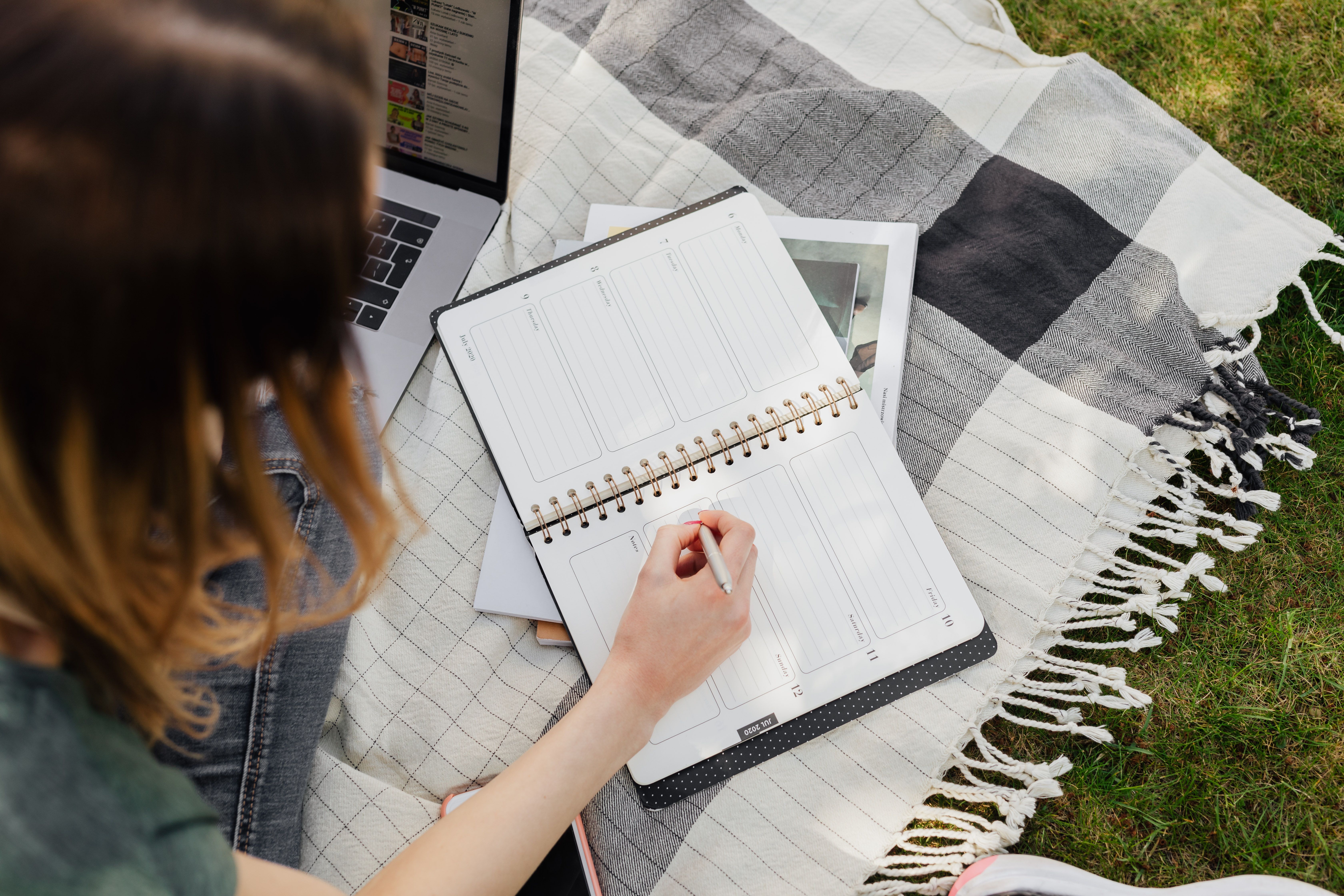 Student writing in organizer while sitting on picnic rug · Free Stock Photo