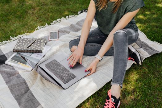 From above of crop anonymous woman in casual clothes searching for information on laptop while sitting on blanket in sunny green park