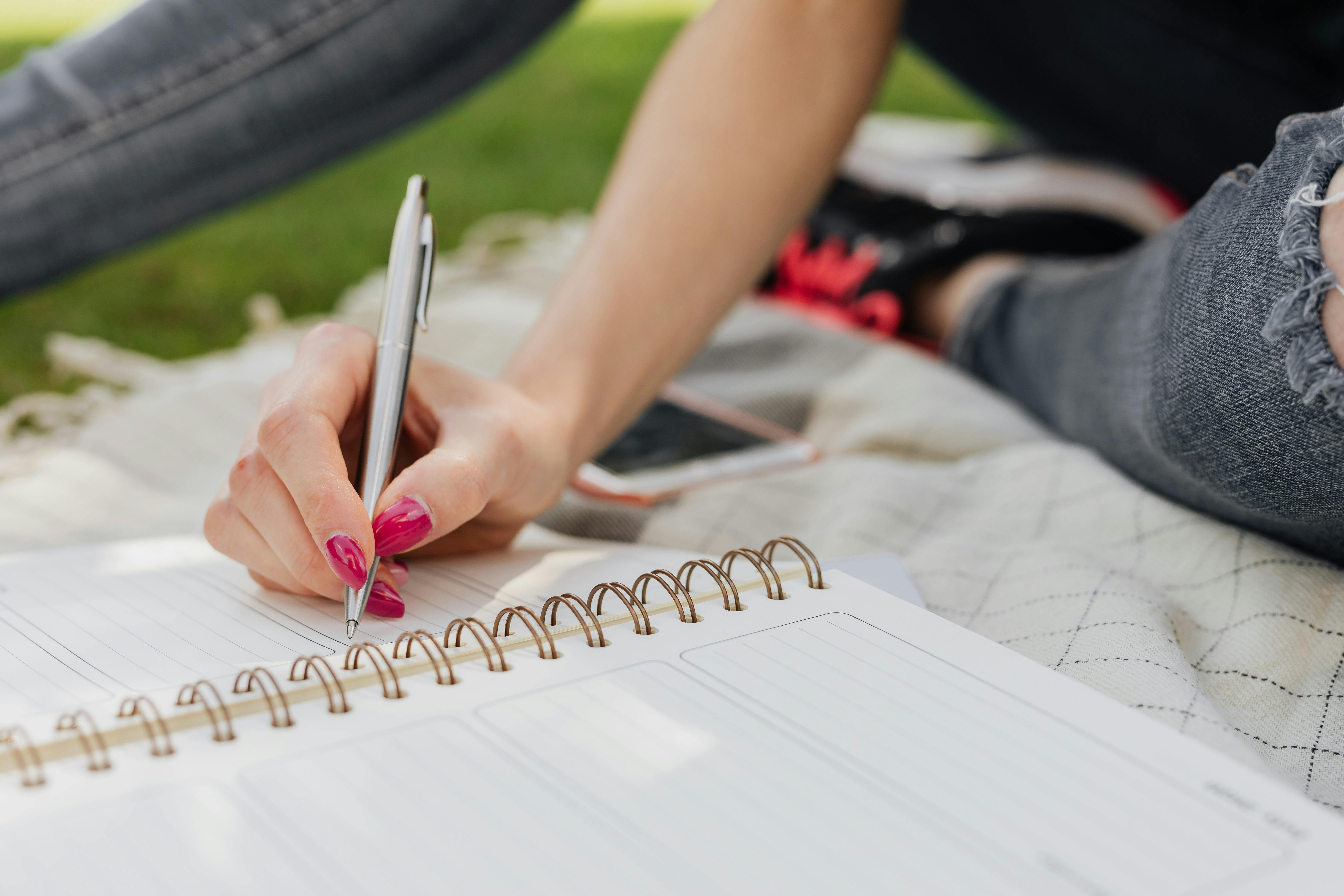 Free Crop anonymous lady in casual wear writing down plans for next day in organizer while sitting on blanket in quiet summer park Stock Photo