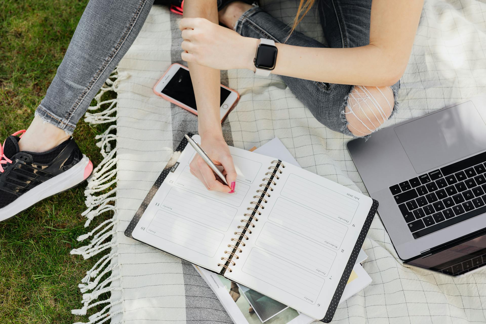 High angle of busy female student writing down plan of studies while sitting with open laptop on blanket in quiet green park