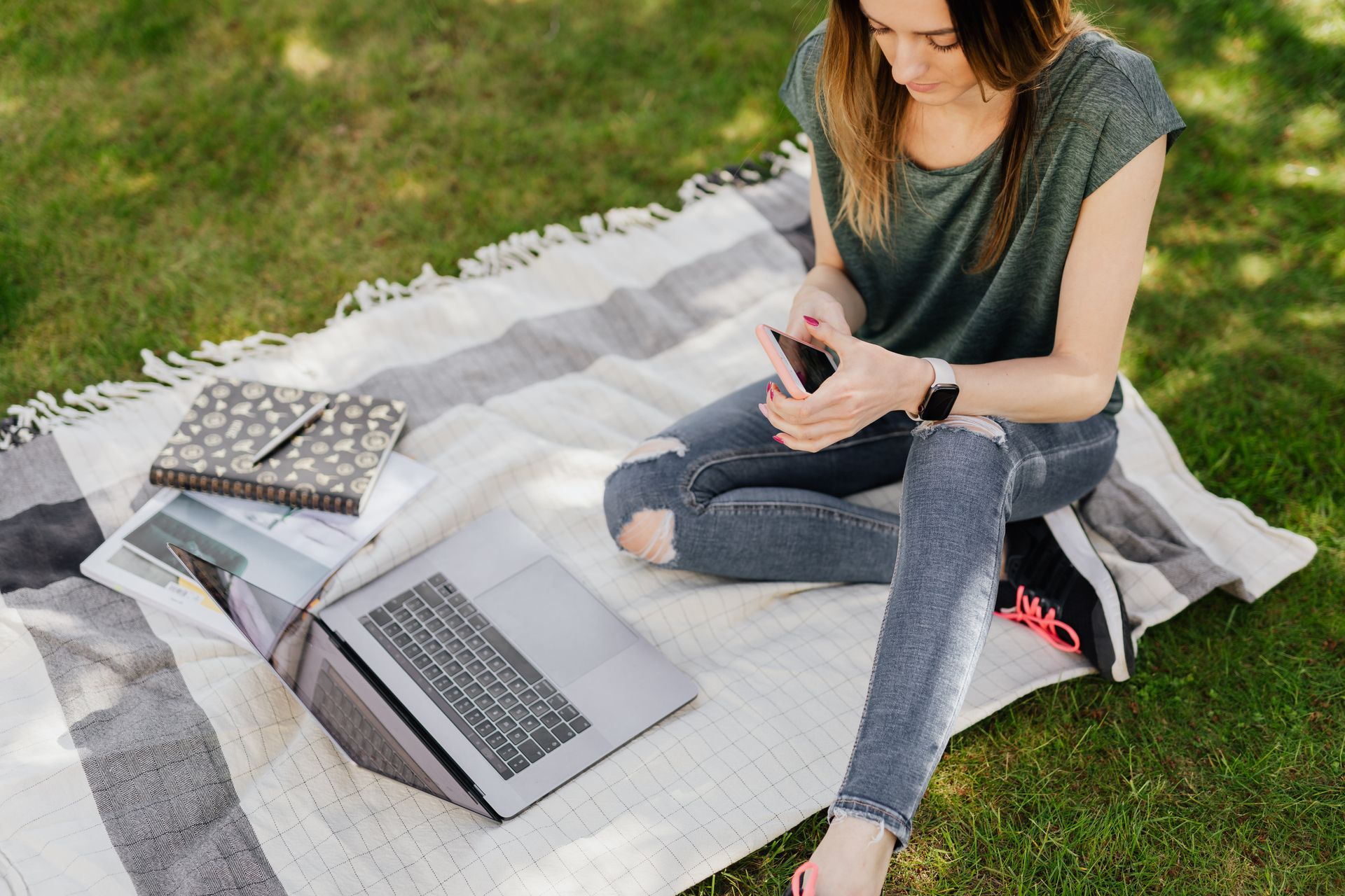 From above of young crop female student in casual clothes surfing social media on smartphone while sitting with laptop and notebooks on blanket placed on green grass in sunny park