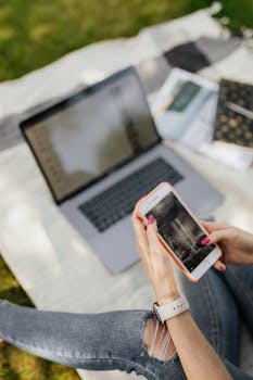 一名女子躺在毯子上，用智能手机发短信，旁边放着笔记本电脑