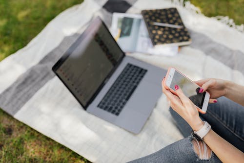 From above crop anonymous female student in blue jeans text messaging on smartphone while sitting on blanket with laptop and college books in green park