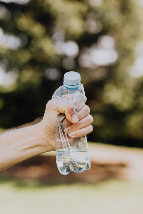 Free Crop unrecognizable man crumpling plastic bottle of water in hand against blurred background of green park trees on sunny summer day Stock Photo