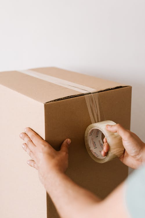 Free Crop anonymous male using tape to seal packed cardboard box against white wall while moving into new place Stock Photo