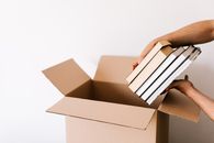 Crop unrecognizable male packing staked books into carton box while standing against white wall during relocation