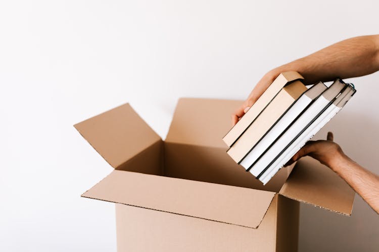 Crop Man Packing Stack Of Books In Opened Cardboard Box