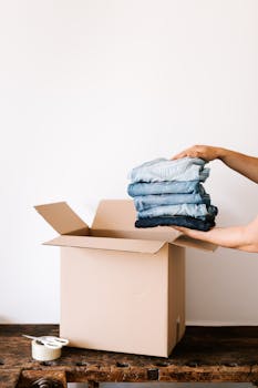 Person packing folded jeans into a cardboard box, preparing for a move or organization task.