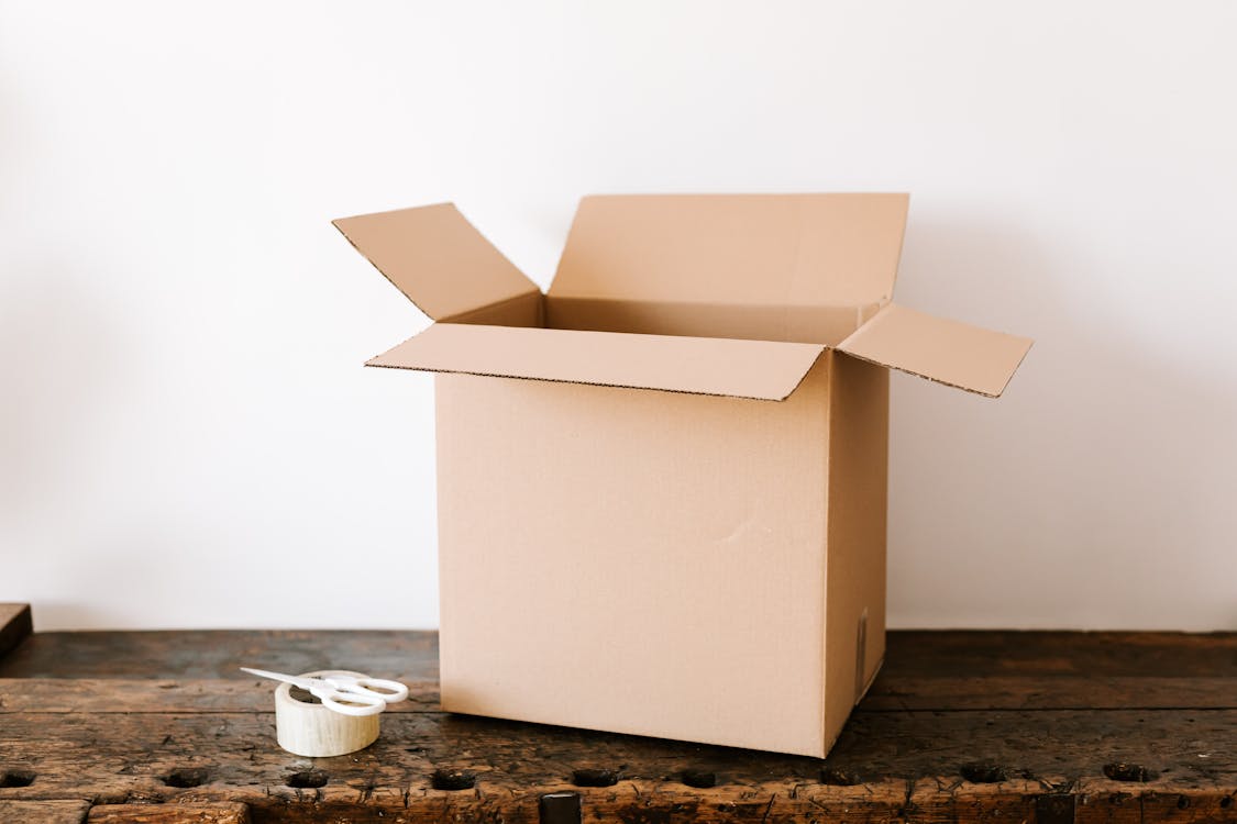 Opened cardboard container placed on old wooden desk near tape and scissors before relocation against white wall