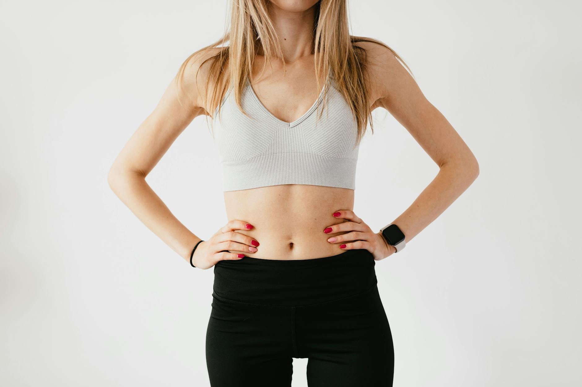 Faceless slim anonymous blond female in sports bra and black leggings in wearable bracelet showing perfect belly on white background while standing with hands on waist