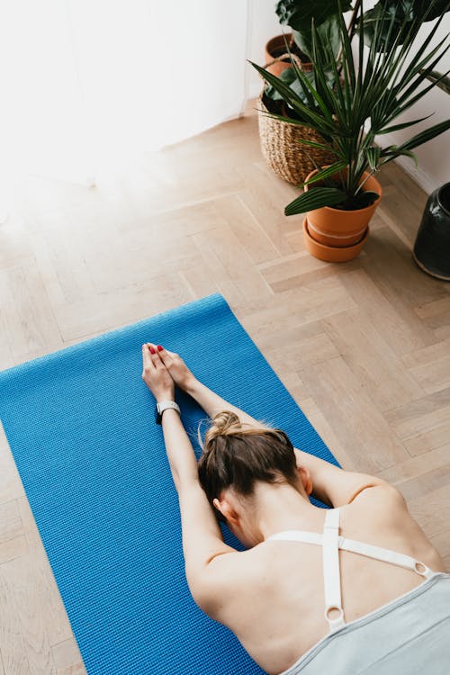 Top view of fit faceless lady in sports clothes lying on blue yoga mat in Child pose at home near potted plants