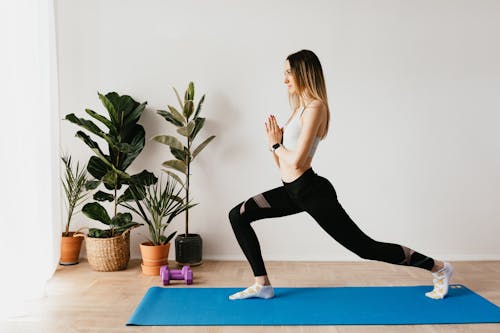 Side view of fit lady in sports clothes and wristwatch standing in Anjaneyasana pose while practicing yoga on mat near dumbbells and potted plants at home and looking away